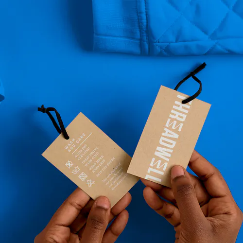 Two hands holding two clothing tags with black ties printed on Kraft paper and with Threadwell in white.