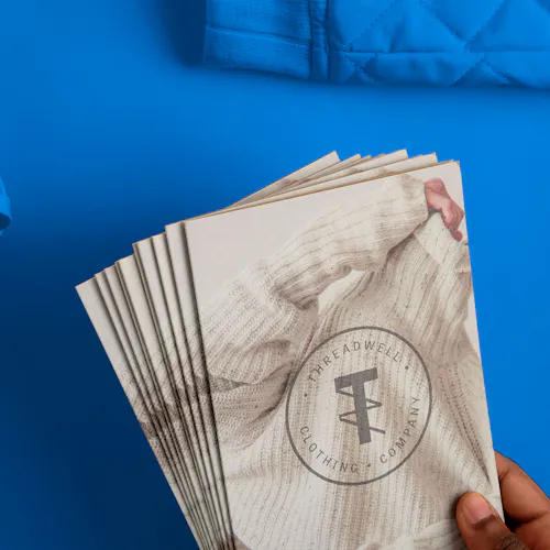 A hand holding fanned-out retail brochures printed with a person in a cream sweater and Threadwell on the cover.