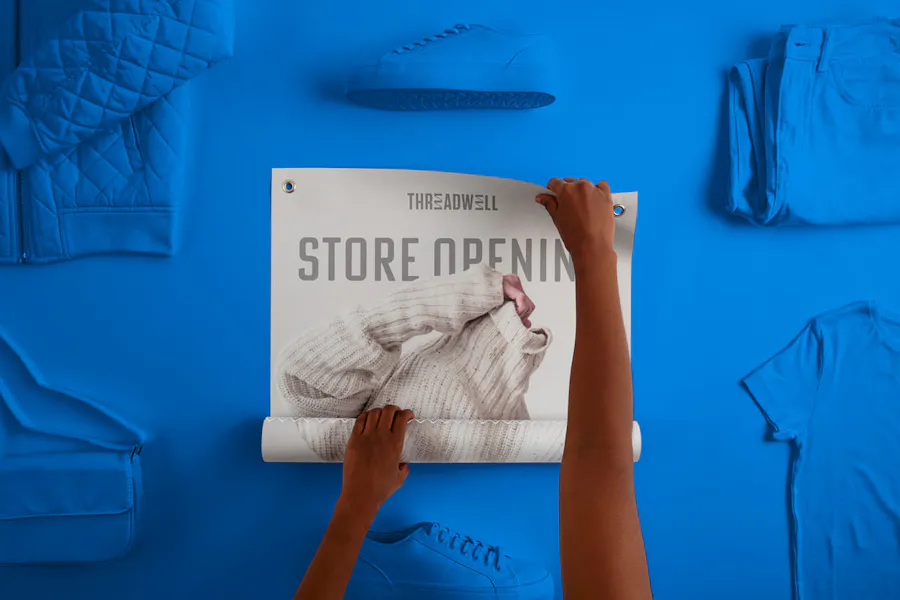 Two hands unrolling a retail poster printed with Threadwell Store Opening surrounded by blue clothing.