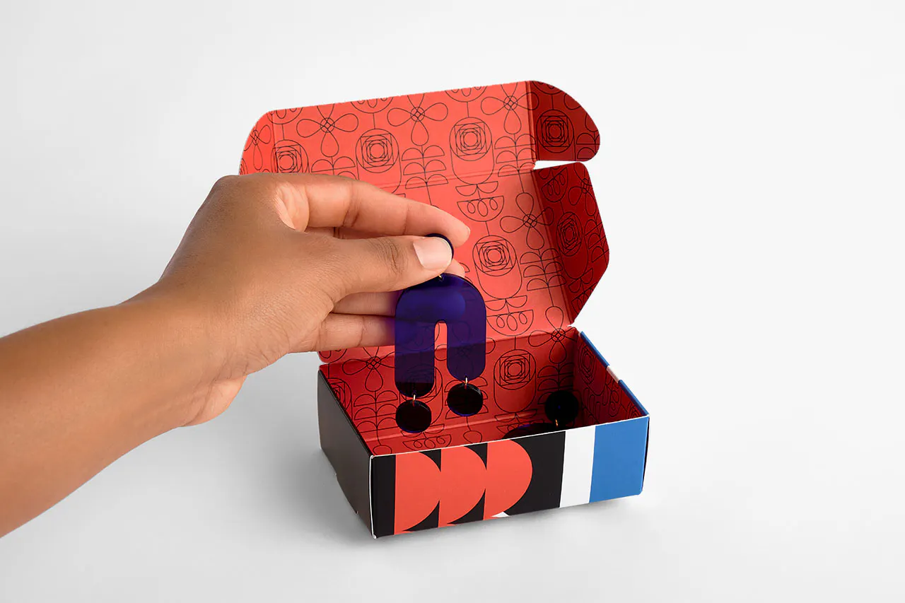 A hand holding a black dangling earring above a custom printed box with the lid open.