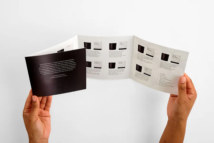 Two hands holding an unfolded brochure with four panels printed with images of candles and product details.