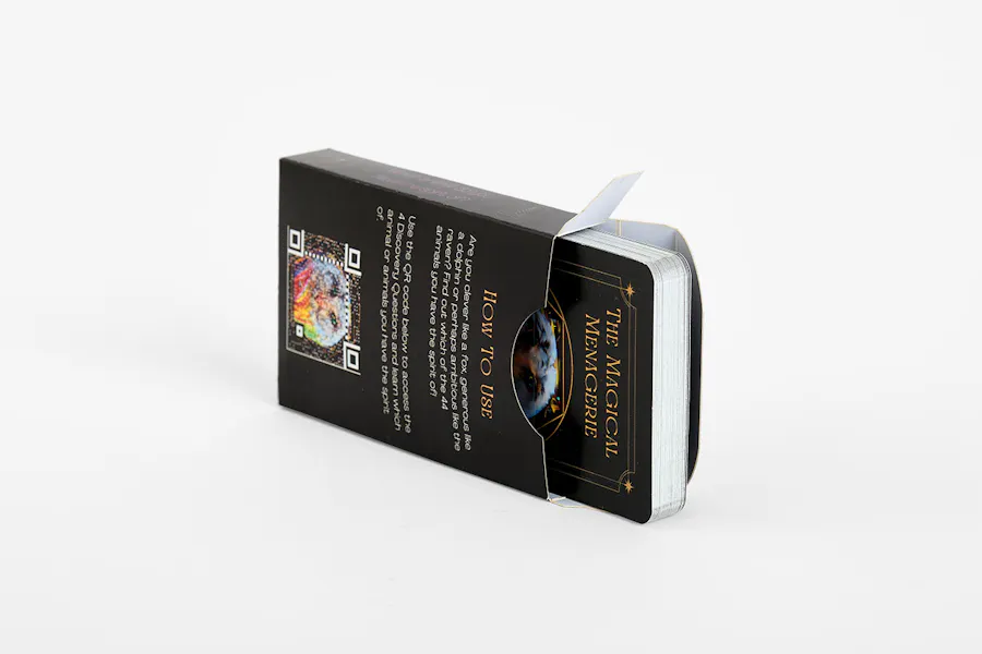 A deck of tarot cards with a black design tipped on its side and coming out of a matching black box with gold text.