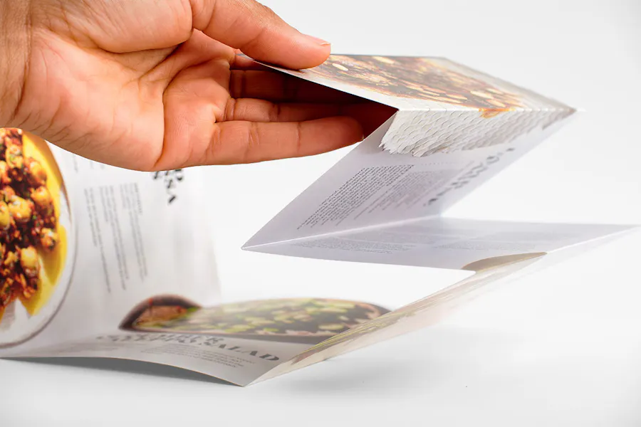 A hand holding up one side of an accordion fold brochure printed with images of food like a salad.