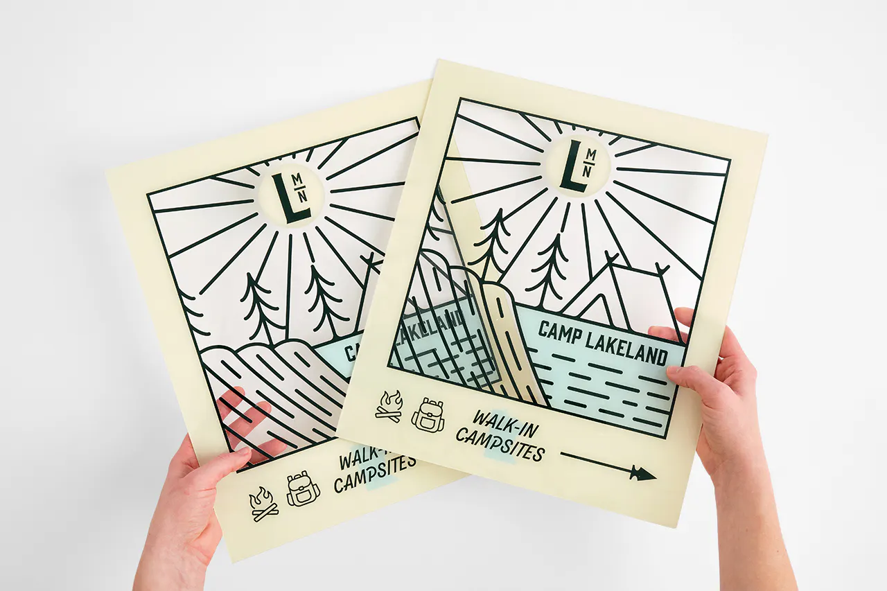 Two hands holding two custom clear signs printed with a camping design and Camp Lakeland.