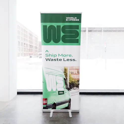 A trade show pop-up banner printed with a green design a Ship More. Waste Less.