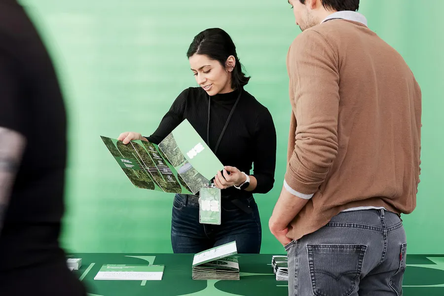 A woman showing a folder to a man standing at a trade show booth with a green background.