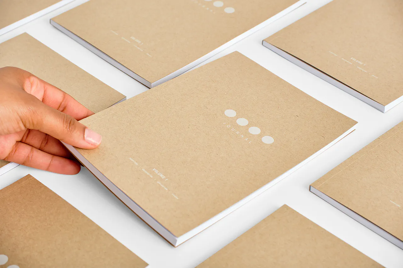 A hand placing a custom notebook with a Kraft paper cover on a table surrounded by more notebooks.