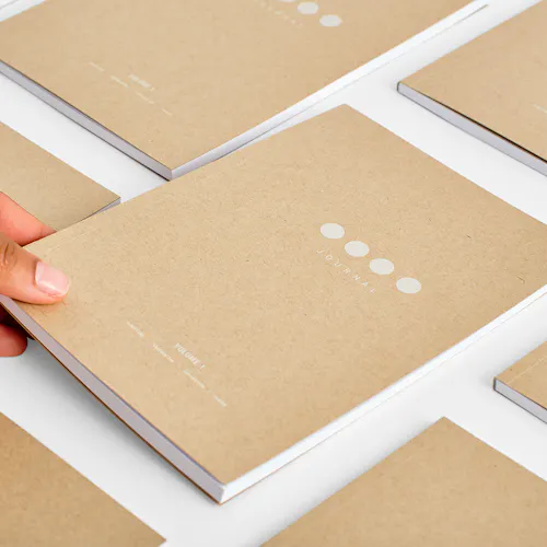 A hand placing a custom notebook with a Kraft paper cover on a table surrounded by more notebooks.