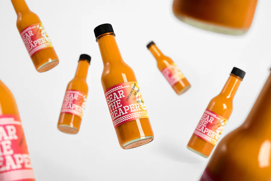Eight hot sauce bottles with black caps and red labels printed with Sear the Reaper.