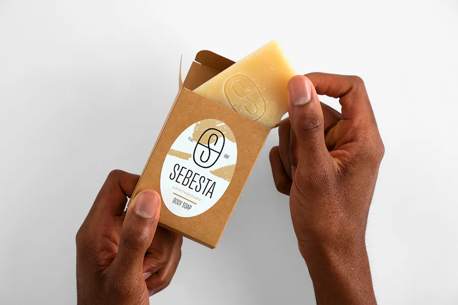 Two hands pulling a bar of soap out of its box with an oval label printed with Sebesta Apothecary.