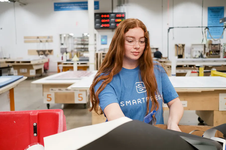 A woman wearing a blue Smartpress T-shirt and safety goggles sorts through recycled magnet material in a production facility.