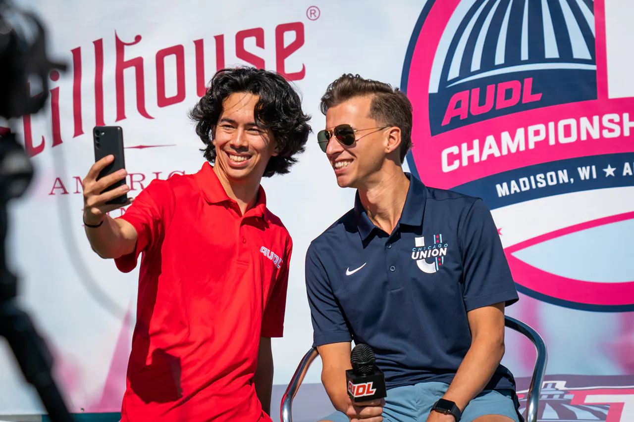 A man in a red polo and a man in a blue polo posing for a selfie in front an AUDL banner.