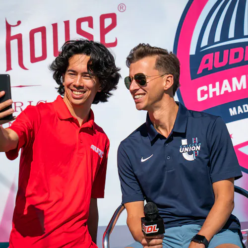 A man in a red polo and a man in a blue polo posing for a selfie in front an AUDL banner.