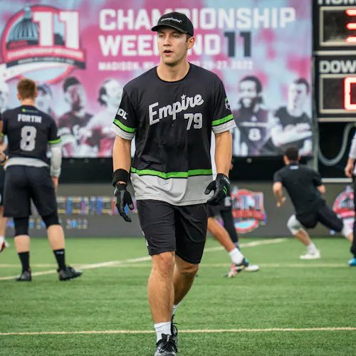 An ultimate disc player wearing a black, white and green jersey walking on the field with a scoreboard behind him.