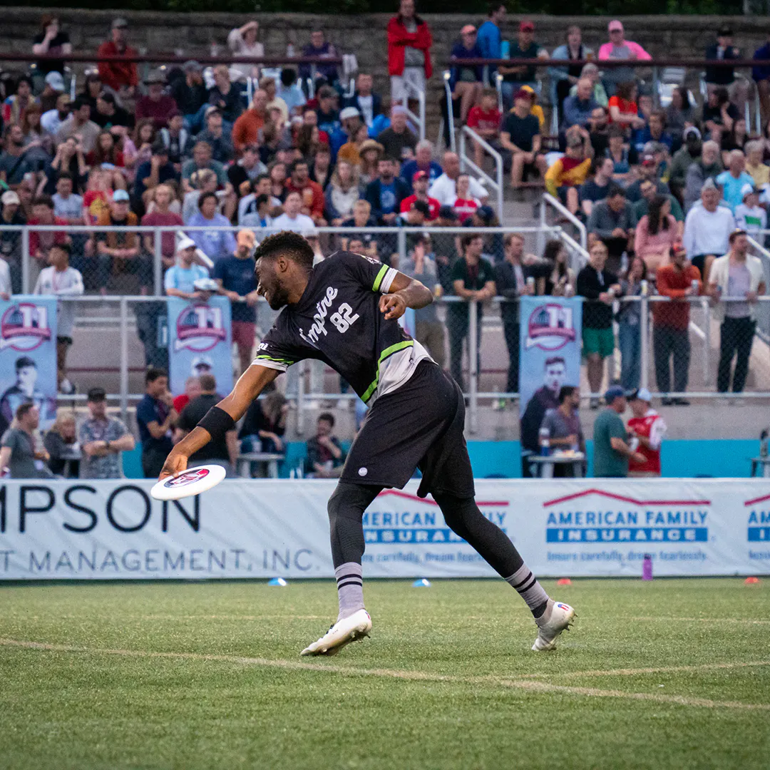 An ultimate disc player in a black, green and white jersey throwing a disc on the field with fans in the crowd in front of him.