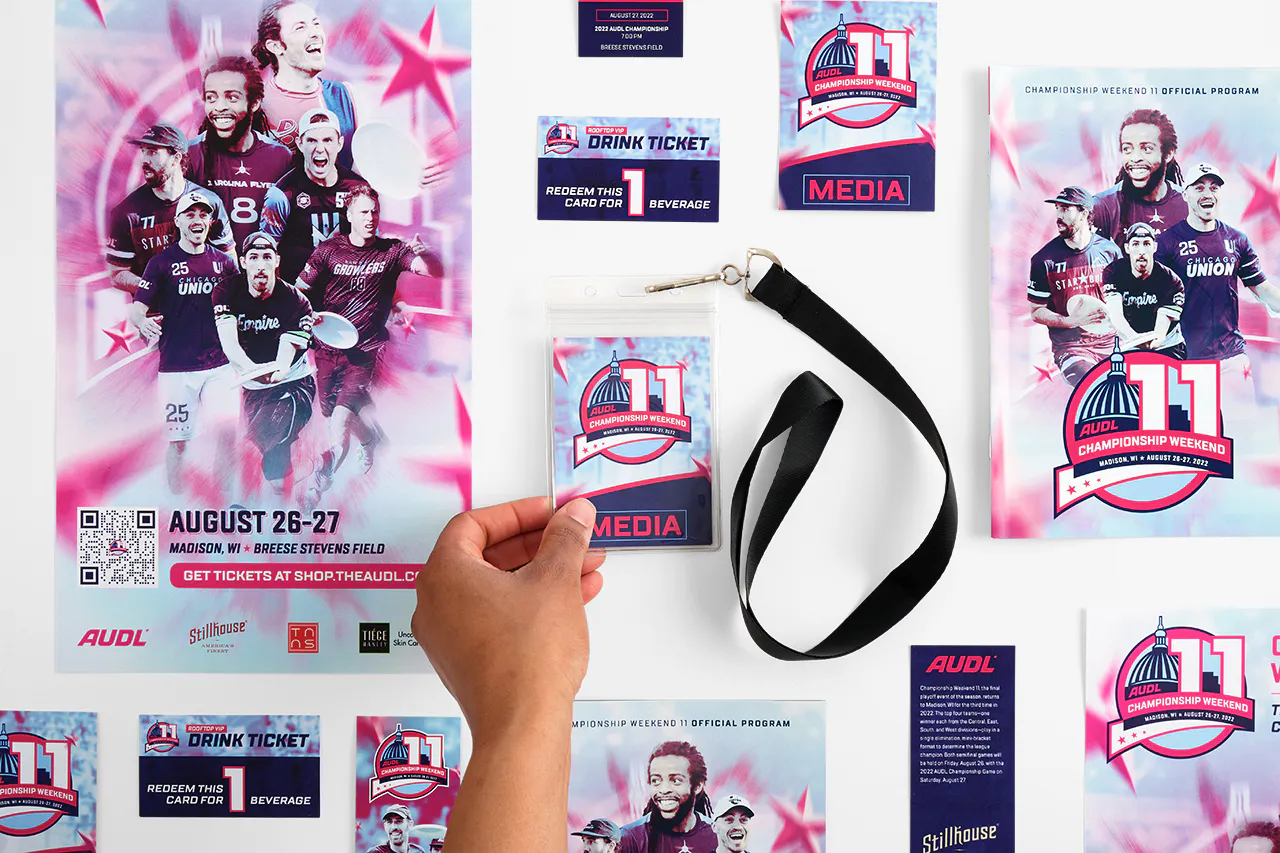 A hand holding a media credential on a black lanyard with posters, event tickets and programs printed for AUDL Championship Weekend 11.