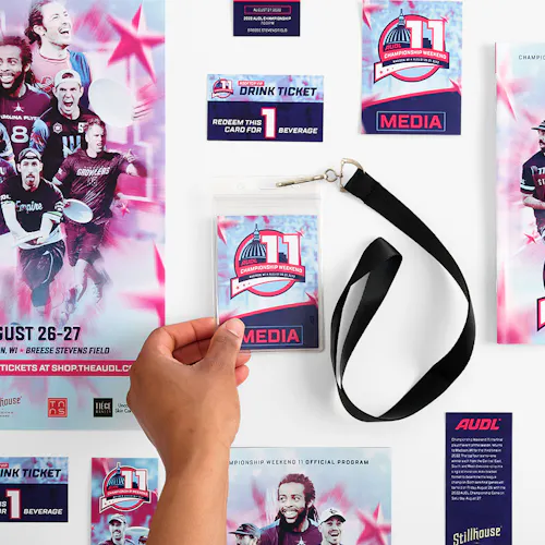A hand holding a media credential on a black lanyard with posters, event tickets and programs printed for AUDL Championship Weekend 11.