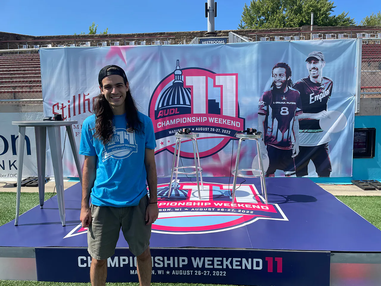 A man with long dark hair smiling and standing in front of a stage and custom banner printed with AUDL Championship Weekend 11.
