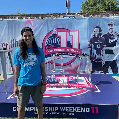 A man with long dark hair smiling and standing in front of a stage and custom banner printed with AUDL Championship Weekend 11.