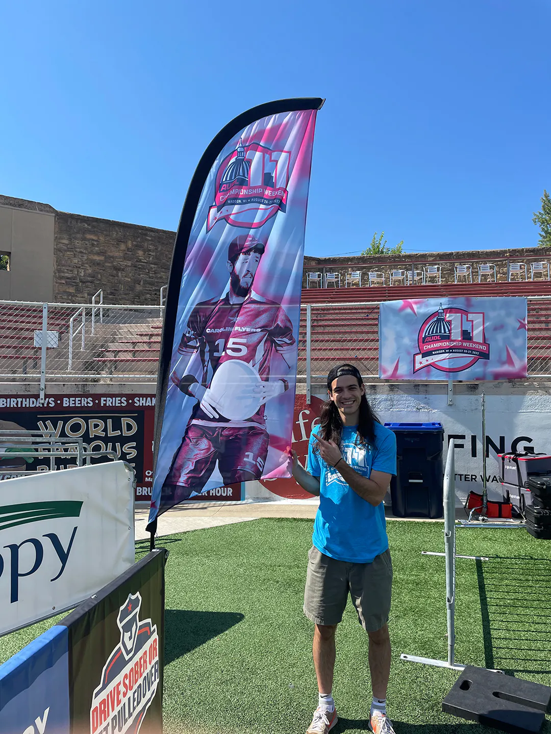 A man smiling and standing next to and pointing at a custom feather flag printed with an image of a man holding an ultimate disc.