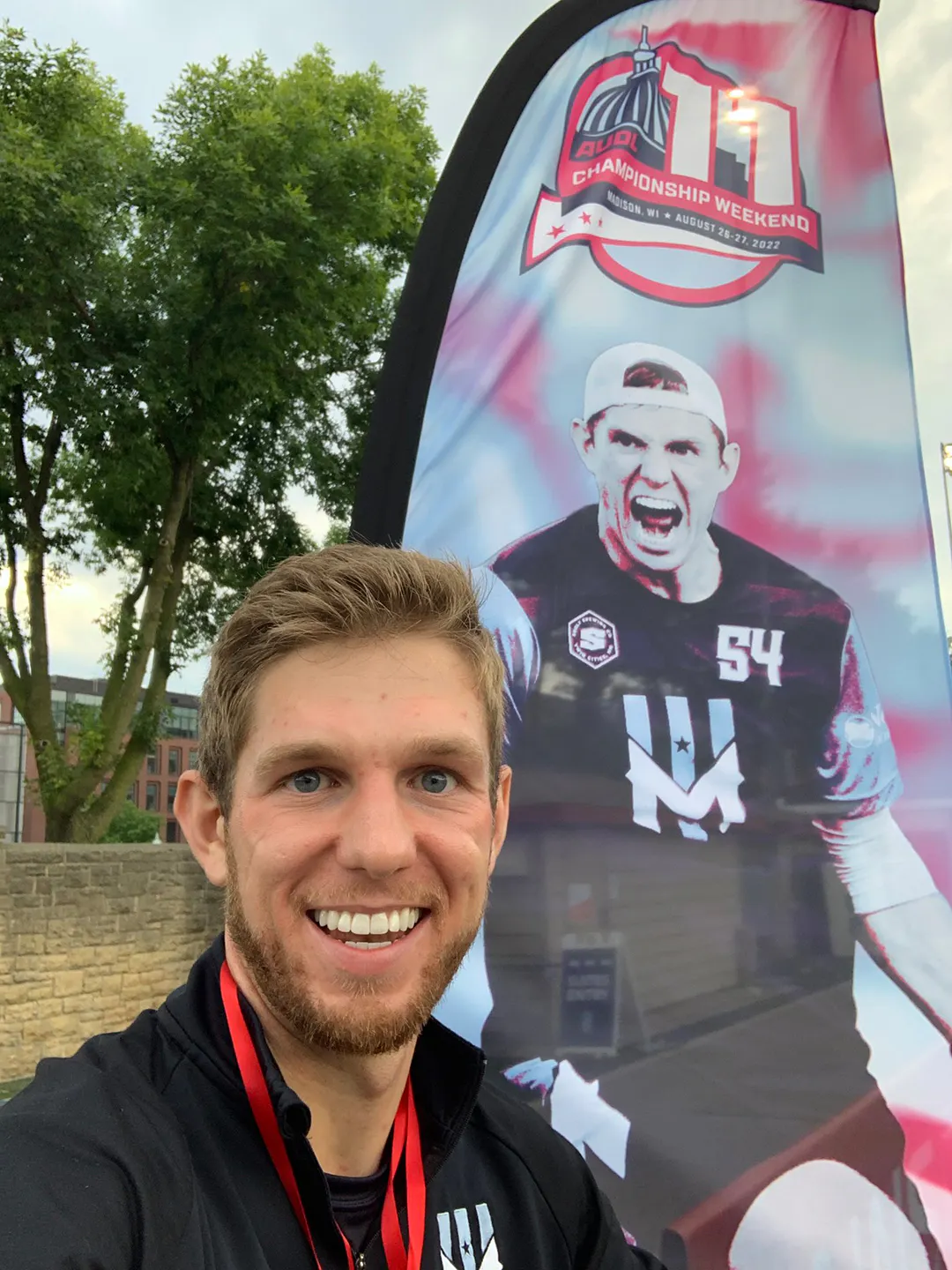 A man smiling and standing in front of a feather flag printed with an image of him and AUDL Championship Weekend 11.