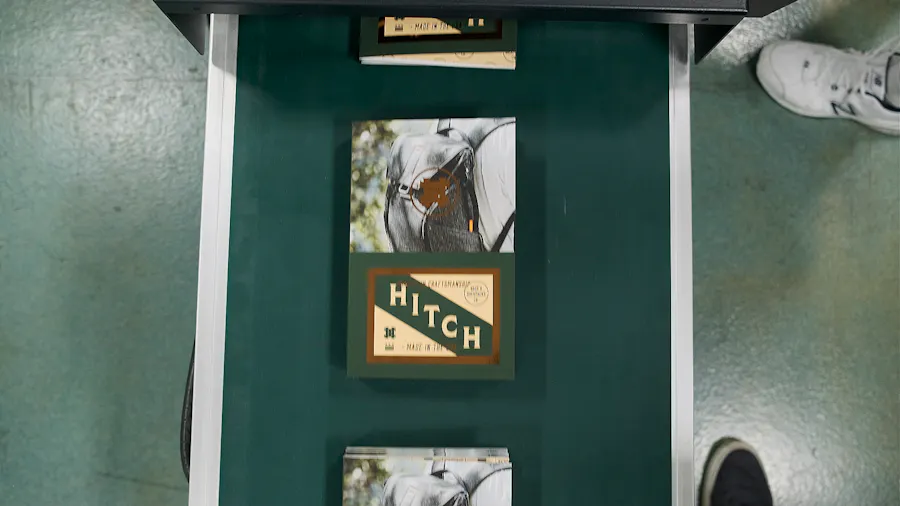 Three custom booklets on a production conveyor belt and printed with a green background, an image of a person wearing a backpack and Hitch.