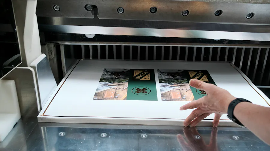 A hand pushing a stack of printed sheets through a guillotine cutting machine in a production facility.