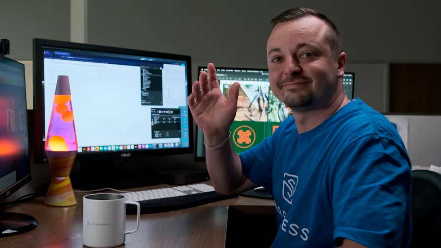 A man in a blue T-shirt waving and sitting at a desk with three computer monitors, a lava lamp and coffee cup in front of him.