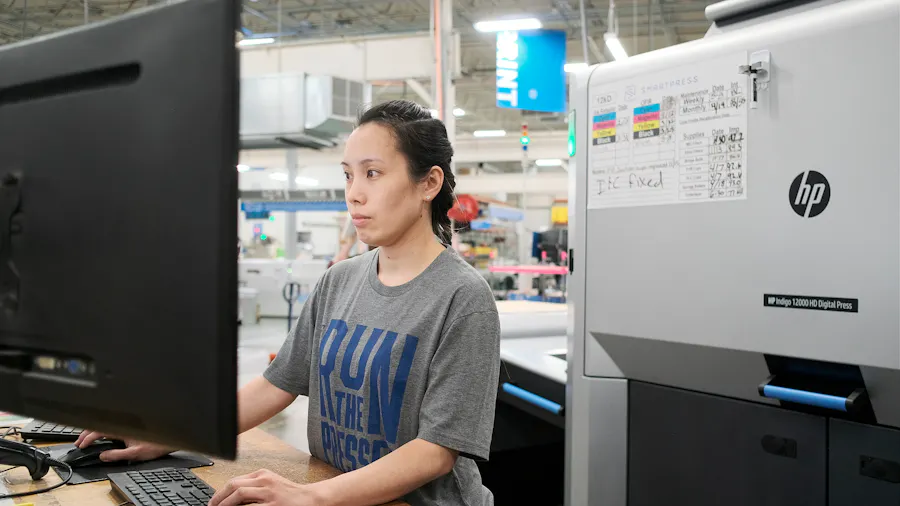 A woman swearing a gray T-shirt and working on a computer at a standing desk next to an HP printer in a production facility.
