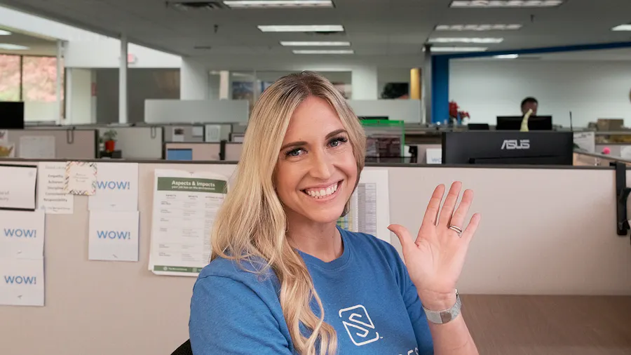 A woman wearing a blue T-shirt sitting at a desk in an office cubicle smiling and waving.