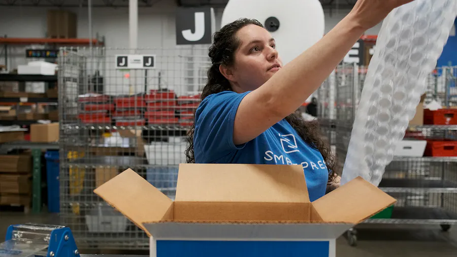 A woman in a blue Smartpress T-shirt holding a sheet of bubble wrap with a box on a table in front of her in a production facility.