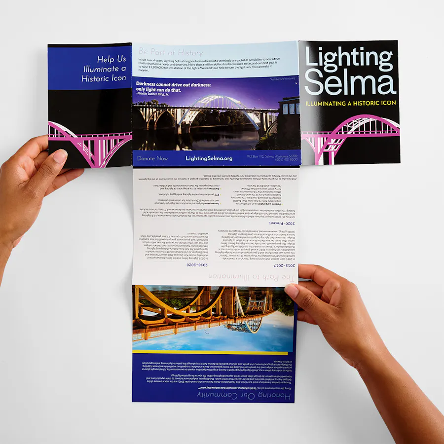 Two hands holding open a T-cross mailer printed with Lighting Selma and images of an illuminated bridge.