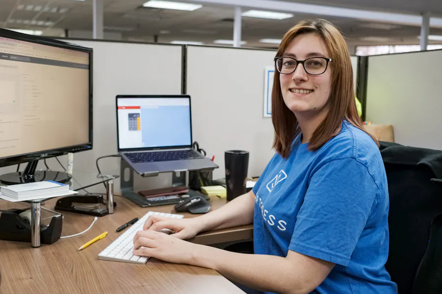 A woman wearing a Smartpress T-shirt sitting at a desk with her hands on a white keyboard and two screens in front of her.