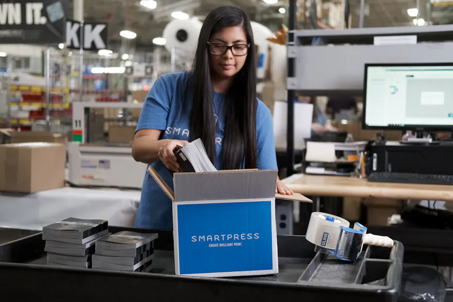 A girl with glasses putting printed booklets into a Smartpress packaging box in a production facility.