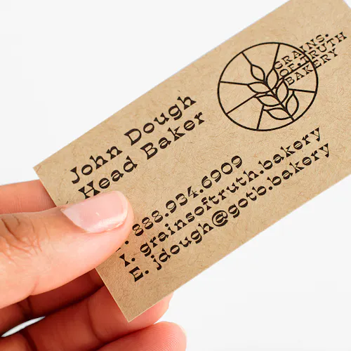 A hand holding a Kraft business card printed with Grains of Truth Bakery and contact info.