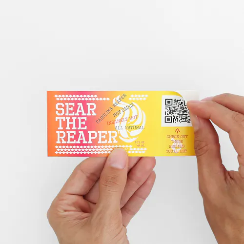 Two hands peeling back the corner of a label printed in orange, pink and yellow with a QR code in the corner.