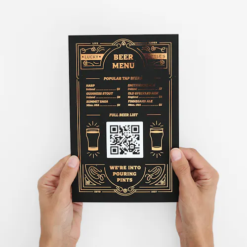 Two hands holding a beer menu printed with rose gold foil, a black background and a QR code in the middle.
