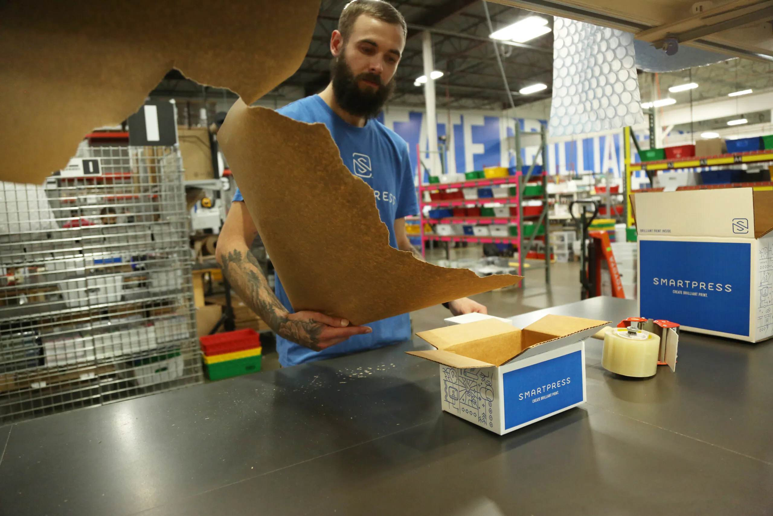 A man in a production facility tearing a piece of Kraft paper and standing at a table with Smartpress boxes on it.
