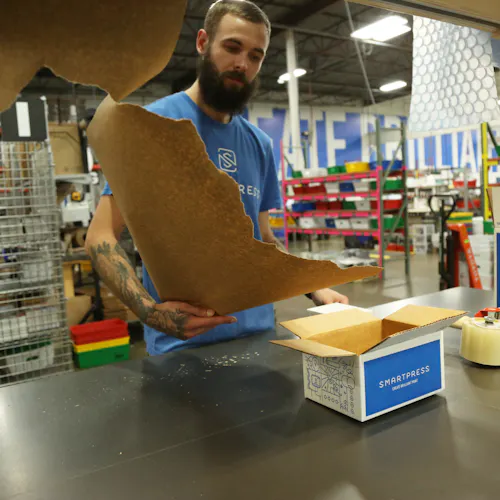 A man in a production facility tearing a piece of Kraft paper and standing at a table with Smartpress boxes on it.