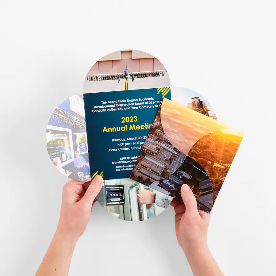 Two hands holding two square petal brochures printed with annual meeting details.