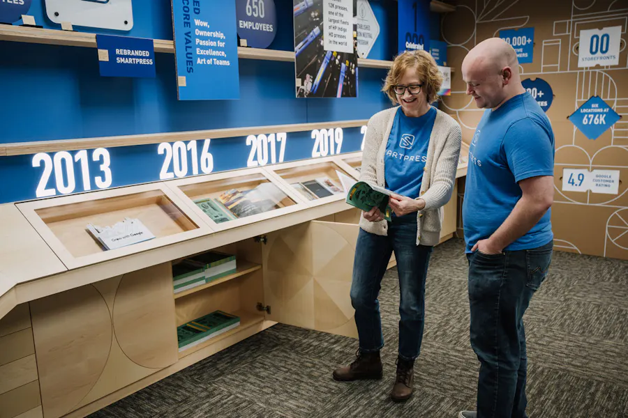 A man and woman wearing Smartpress T-shirts flipping through a booklet by a timeline wall.