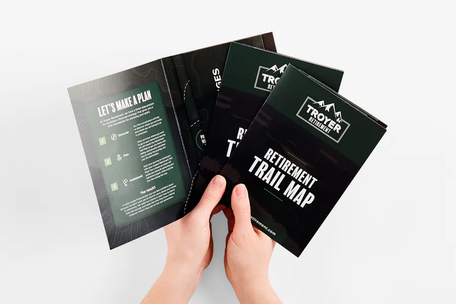 Two hands holding three fanned-out brochures printed with a black and green design and retirement information.