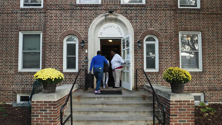 A woman holding open the door of a brick building for two people and a St. Anne's Place sign above the door.