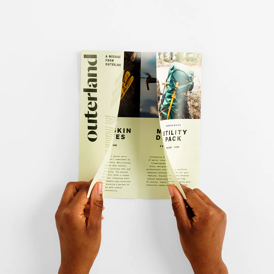 Two hands unfolding a closed gate brochure printed with Outerland and outdoor gear details.