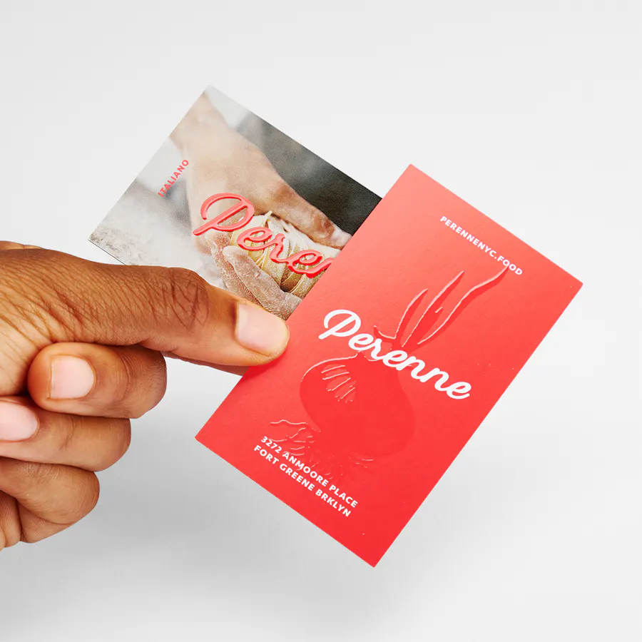 A hand holding two raised UV business cards printed with Perenne, a red background and image of handmade pasta.