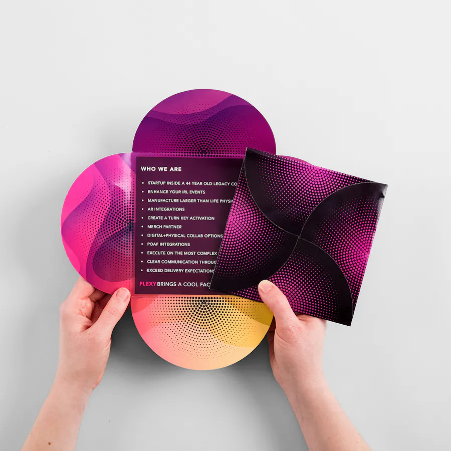 Two hands holding a folded and unfolded square petal brochure printed with a magenta, orange and black design.