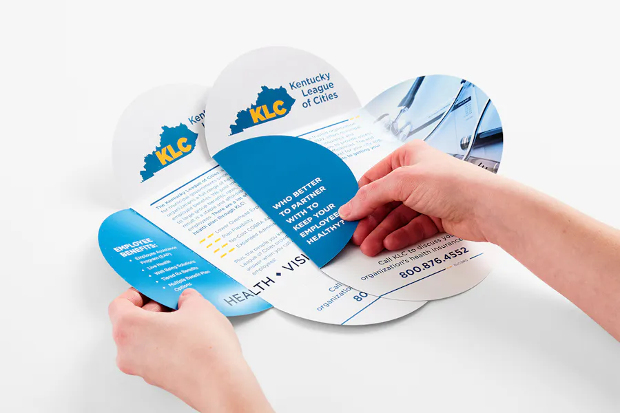 Two hands holding two unfolded sales brochures printed with a petal fold and health insurance info.