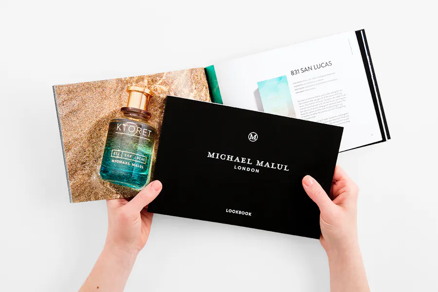 Two hands holding two brand lookbooks printed with images of fragrance bottles and Michael Malul in white.