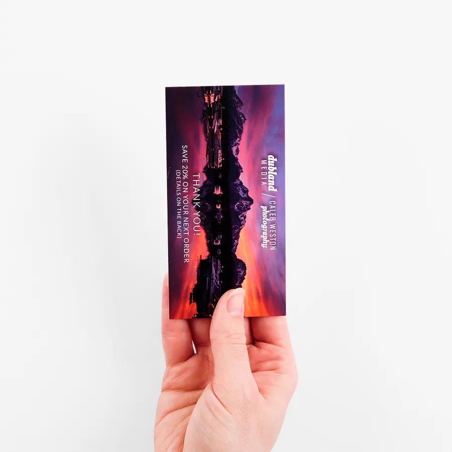 A hand holding a custom bookmark printed with an image of a mountain and a purple, orange and pink sunset.