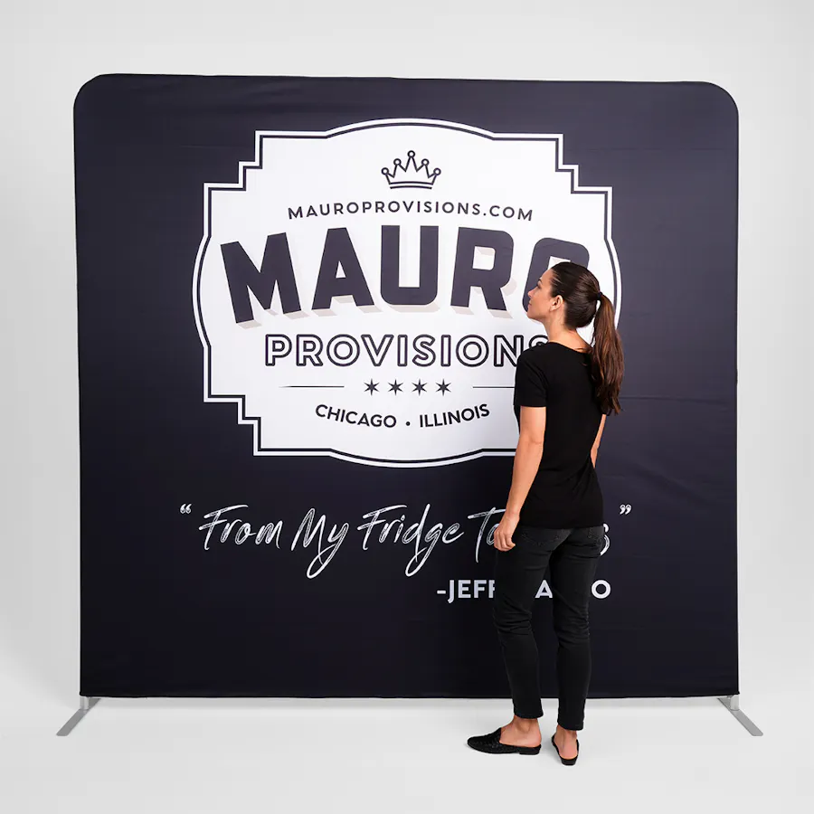 A woman in a black outfit standing in front a tension fabric display printed with Mauro Provisions in black and white.